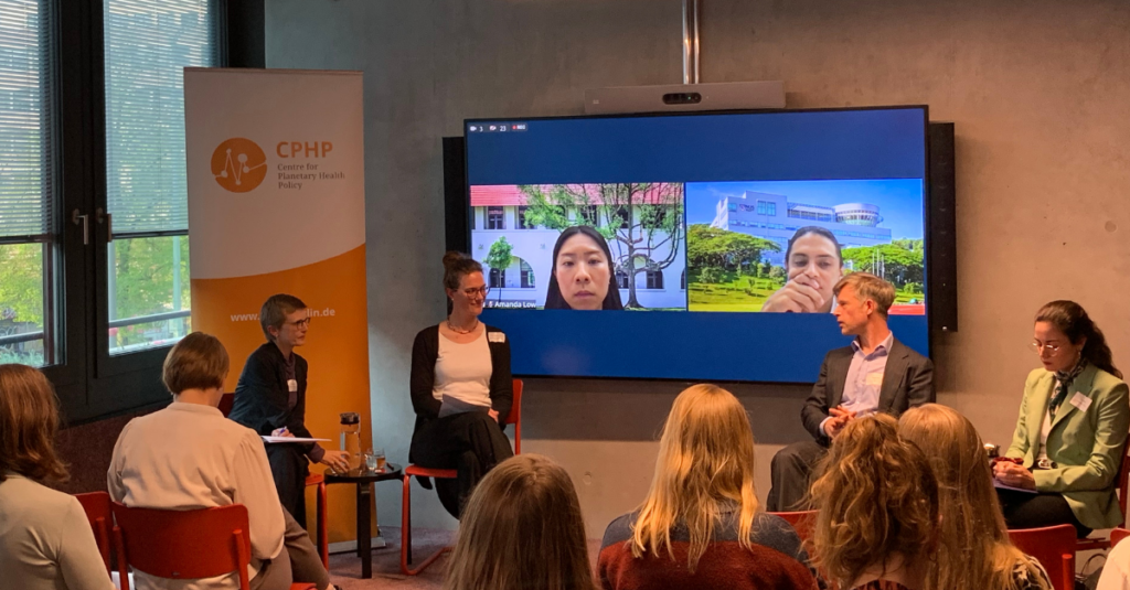 Die Moderatorin und die drei Referent*innen sitzen in einem Raum vor einem Bildschirm mit unscharfen Gesichtern, mehrere weitere Personen sitzen im Publikum, im Hintergrund ein Banner mit dem Logo und Schriftzug 'CPHP'