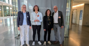 Vier Personen stehen nebeneinander vor einer Glaswand mit dem Schriftzug 'Deutscher Bundestag'
