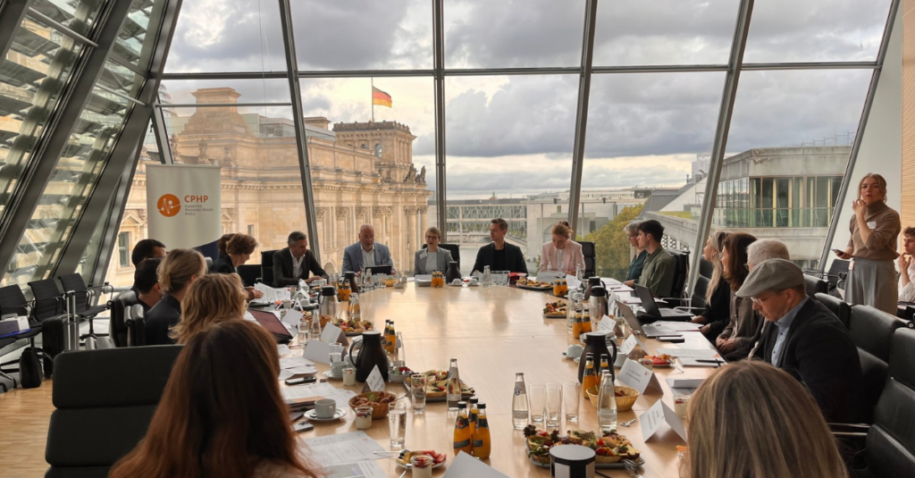 Menschen sitzen an einem großen Tisch in einem Raum mit Glasfassade und Blick auf das Reichstagsgebäude.